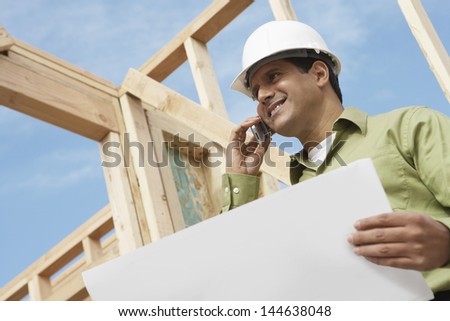 Low angle view of a smiling construction worker with cellphone and blueprints at site