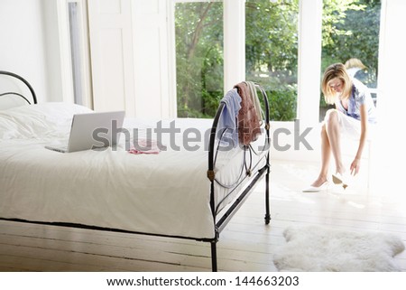 Young woman putting on shoes with laptop on bed