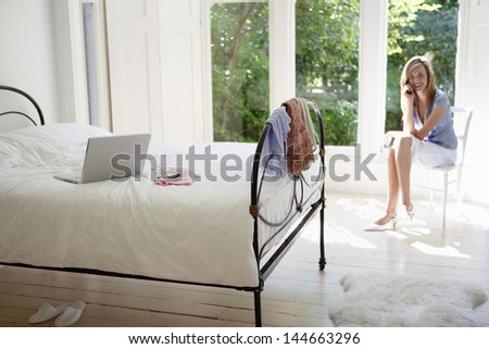 Young woman using cell phone with laptop on bed