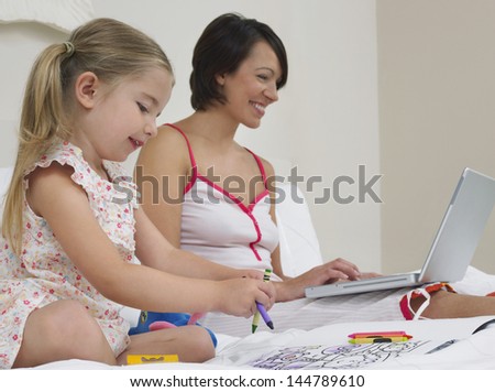 Mother using laptop by daughter as she draws in bed
