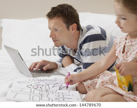 Father using laptop by daughter as she draws in bed