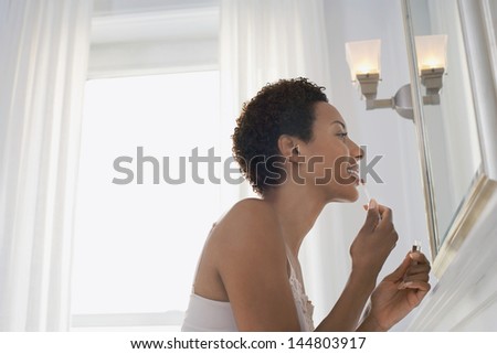 Side view of an African American woman applying lip gloss in mirror at home