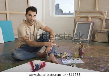Portrait of a male artist sitting with painting tools on floor at a studio