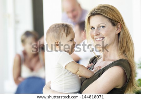 Portrait of young mother carrying baby on porch with family behind