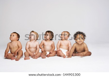 Row of multiethnic babies sitting side by side looking away isolated on gray background Row of multiethnic babies sitting side by side looking away isolated on gray background