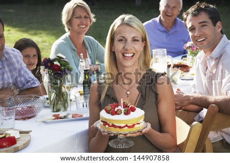 Portrait of cheerful young woman holding birthday cake with family sitting at dining table in garden