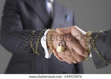 Midsection of two businessmen shaking hands wrapped in gold chain and padlock against gray background