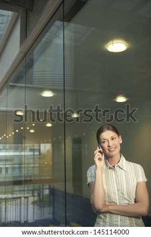 Young businesswoman using mobile phone while standing against glass wall in office