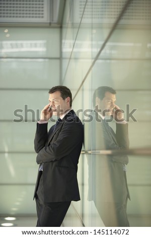 Side view of middle aged businessman using cellphone while leaning on glass wall in office