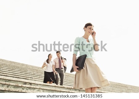 Happy businesswoman using mobile phone on steps with colleagues in background against clear sky