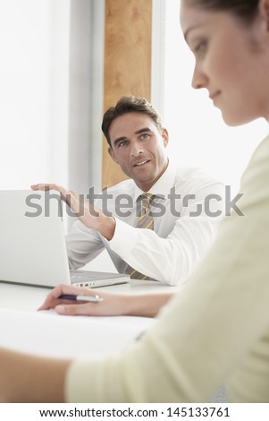 Young businessman with laptop looking at female colleague during meeting in office