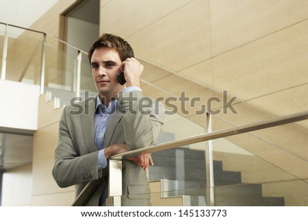 Young businessman using cellphone while leaning on glass railing in office