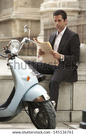 Young businessman sitting by scooter reading newspaper