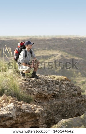 Full length of young man with backpack crouching on top of mountain