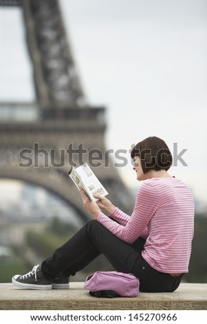 Side view of a young woman sitting on balcony and reading book in front of Eiffel Tower