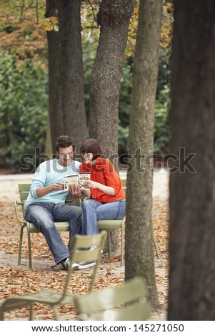 Young couple reading guidebook on park chairs along tree trunks