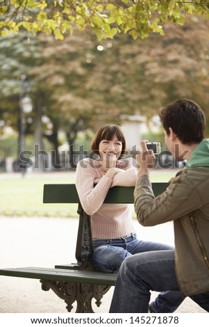 Cropped man photographing smiling young woman on park bench