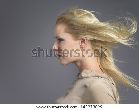 Closeup side view of a young woman with blond hair blowing in wind against gray background