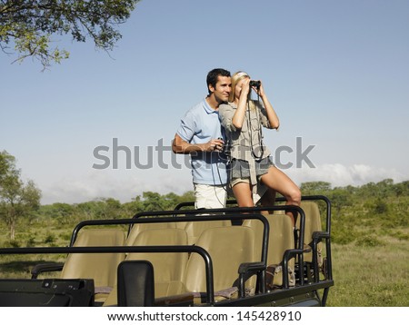 Young Couple On Safari Standing In Jeep And Looking Through Binoculars