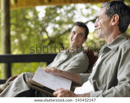 Side View Of A Happy Man With Book Sitting On Terrace With Blurred Male Friend In Background