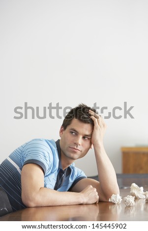 Frustrated young man sitting at conference table with crumpled papers and looking away