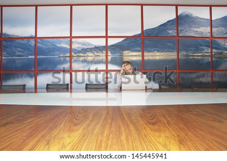 Bored businessman sitting with head in hands using laptop in modern conference room