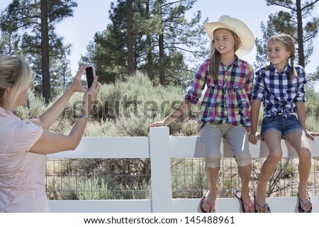 Mother photographing daughters through cell phone as they sit on fence at park