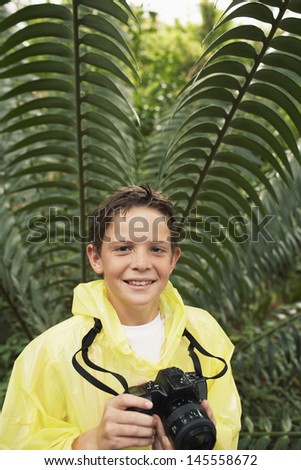 Portrait of happy young boy with camera by large fern in forest during field trip