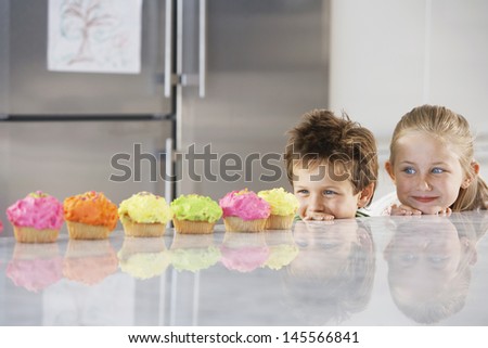 Young Girl And Boy Peeking Over Counter At Row Of Cupcakes
