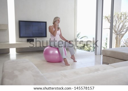 Full length of middle aged woman sitting on fitness ball while using cell phone in living room