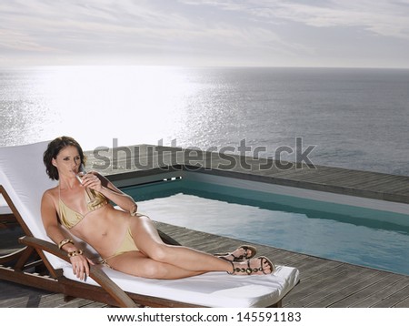 Young woman in bikini lying on deck chair sipping champagne at poolside by ocean