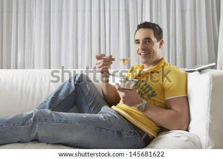Portrait of a young man sitting on sofa and eating noodles from takeaway tray