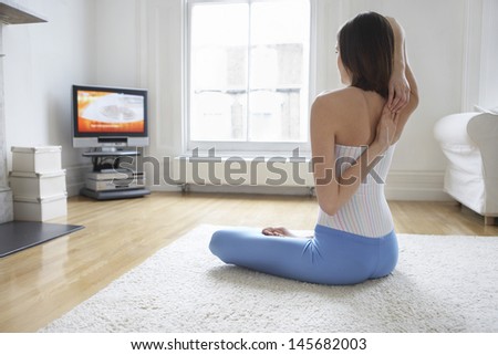 Rear view of a young woman stretching arms on floor and watching television at home