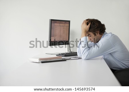 Side view of a young businessman with head in hands by computer sitting at office desk