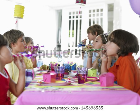 Side view of six children at outdoor table blowing party puffers