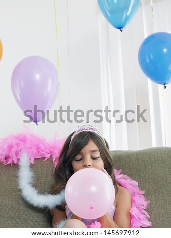 Little young girl sitting on sofa and blowing up balloon