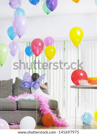 Rear view of a young girl in fairy wings on sofa amid balloons at home