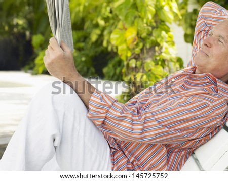 Side view of a relaxed mature man reclining on lounge chair and reading newspaper