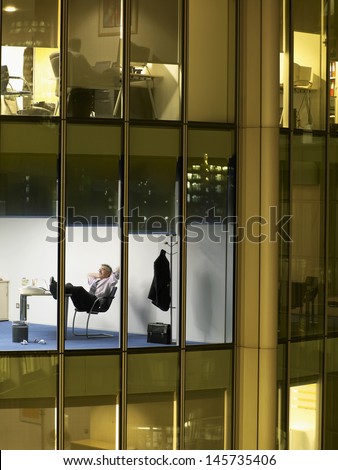 View of middle aged businessman relaxing with feet on desk at night through window