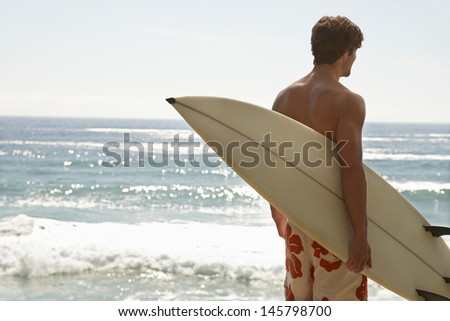 Rear view of young man carrying surfboard looking at ocean