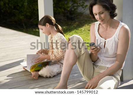 Woman using cell phone with daughter reading book on porch