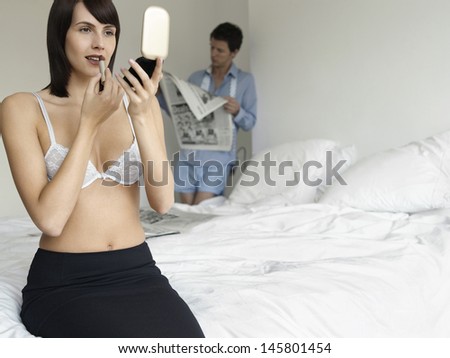 Semi dressed woman applying lipstick on bed with man reading newspaper in background