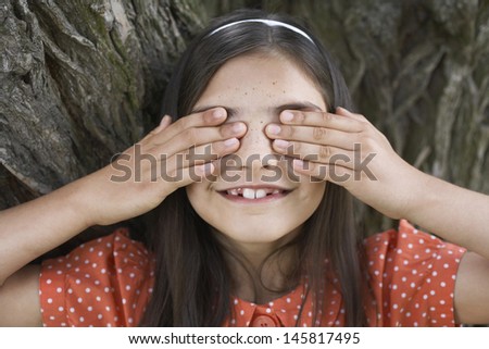 Closeup of happy girl playing hide and seek while covering eyes by tree