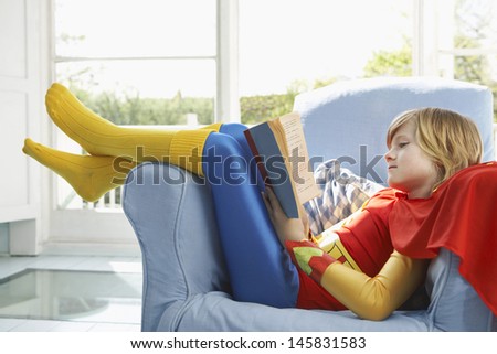 Side view of a relaxed young boy in superhero costume reading book on armchair