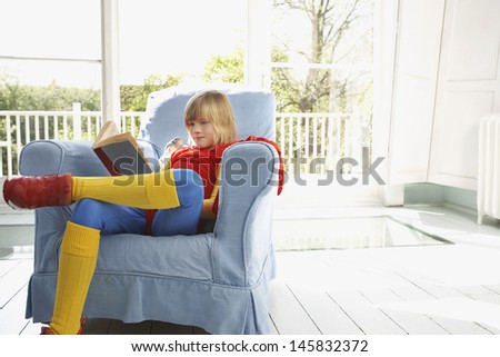 Full length of a blond young boy in superhero costume reading book in armchair