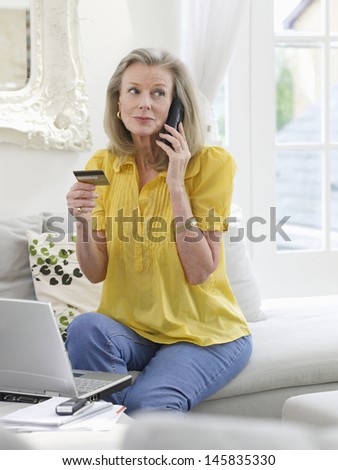 Mature woman with laptop using credit card and phone in living room at home
