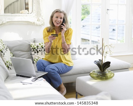 Happy mature woman with laptop using credit card and phone in living room at home
