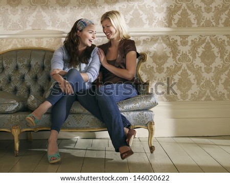 Full length of two cheerful young women sitting on sofa against wallpaper