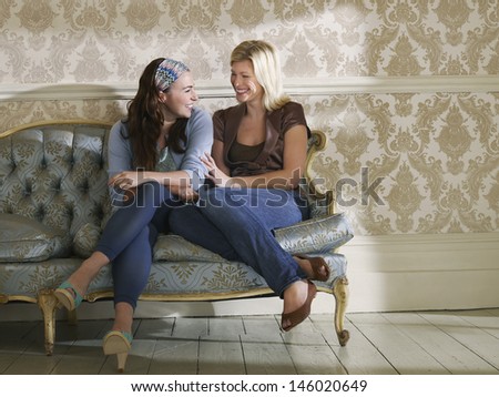 Full length of two cheerful young women sitting on sofa against wallpaper