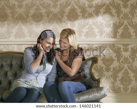 Two smiling young women sitting on sofa against wallpaper